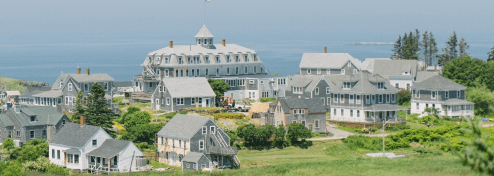 Coastal village with gray houses, greenery, and ocean in the background.