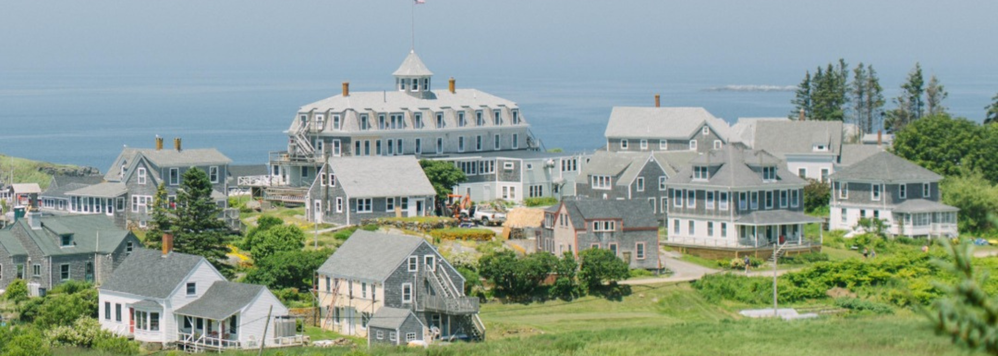 Coastal village with gray houses, greenery, and ocean in the background.